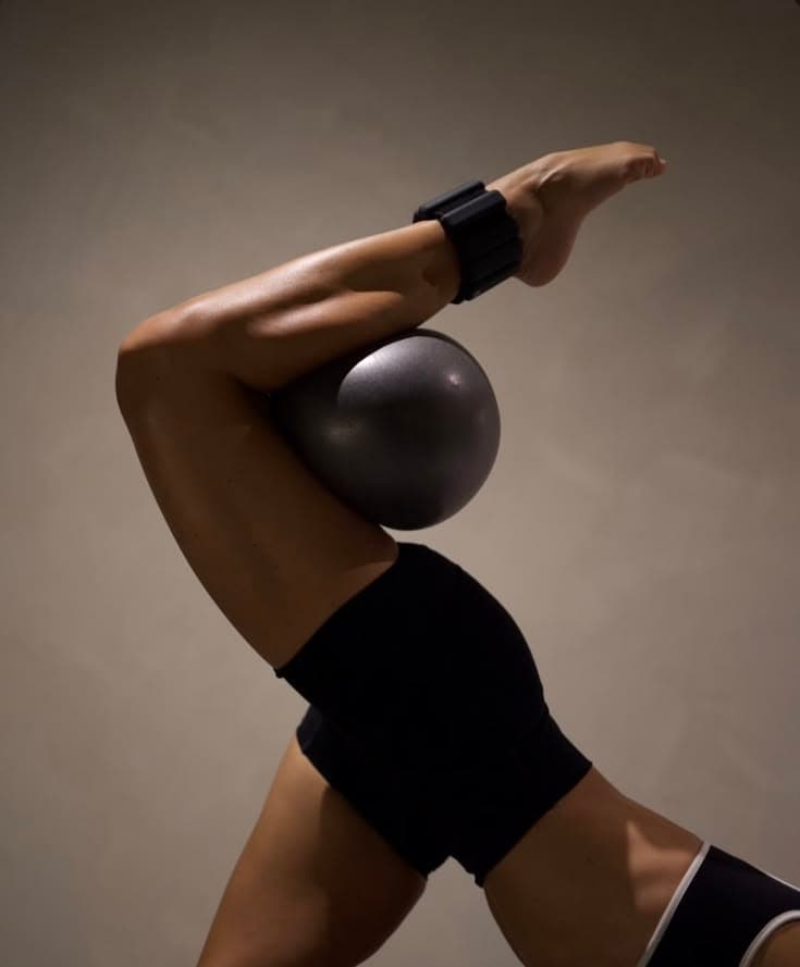 Sculptural crop of a posed figure balancing a Pilates ball against forearm and shoulder